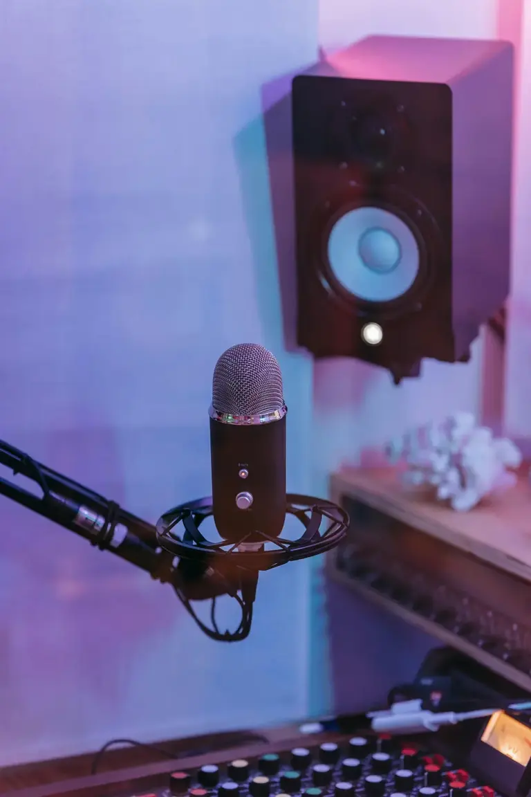 A close-up of a black condenser microphone, with a studio monitor speaker and mixing console in the background.