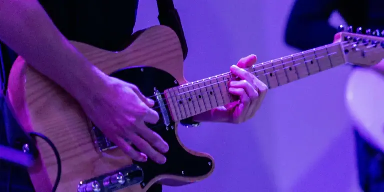 A close-up of a musician's hands playing an electric guitar with a black pickguard under vibrant purple stage lighting.